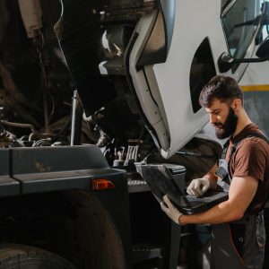 Auto mechanic working on a laptop while diagnosing engine issues in a truck at a busy garage, focusing on efficient troubleshooting