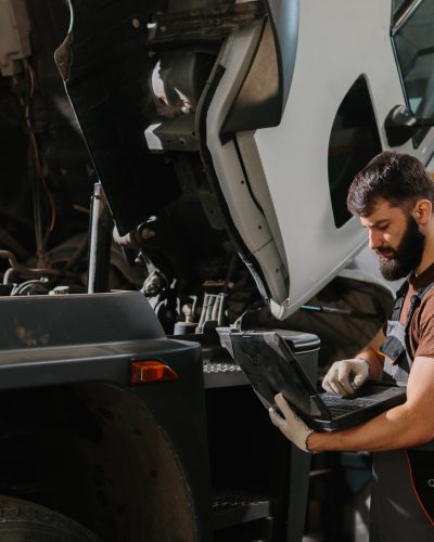 Auto mechanic working on a laptop while diagnosing engine issues in a truck at a busy garage, focusing on efficient troubleshooting